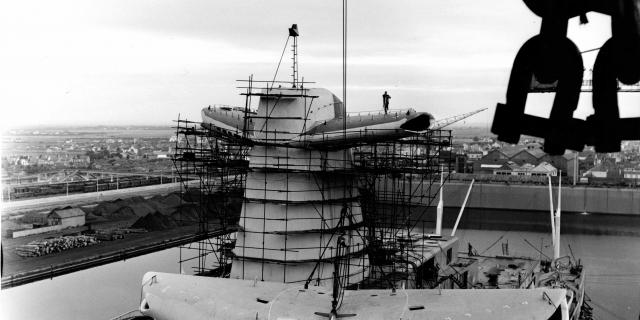Dans le quai d'armement du bassin de Penhoët à Saint-Nazaire, des ouvriers des Chantiers de l’Atlantique procèdent à l'embarquement d'un aileron de cheminée du paquebot France (1962).